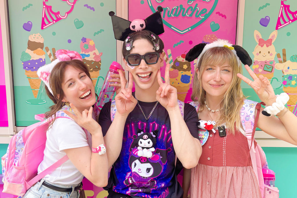 Three people posing in front of a colorful background with kawaii characters and text.
